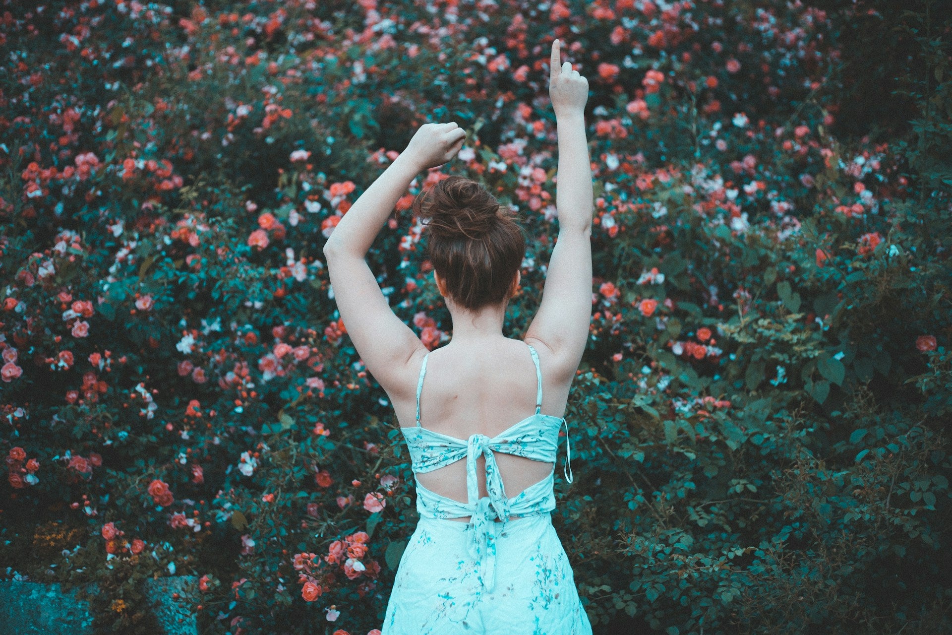 A red-haired woman in a blue floral sundress faces away from the camera, toward a flower-covered bush.