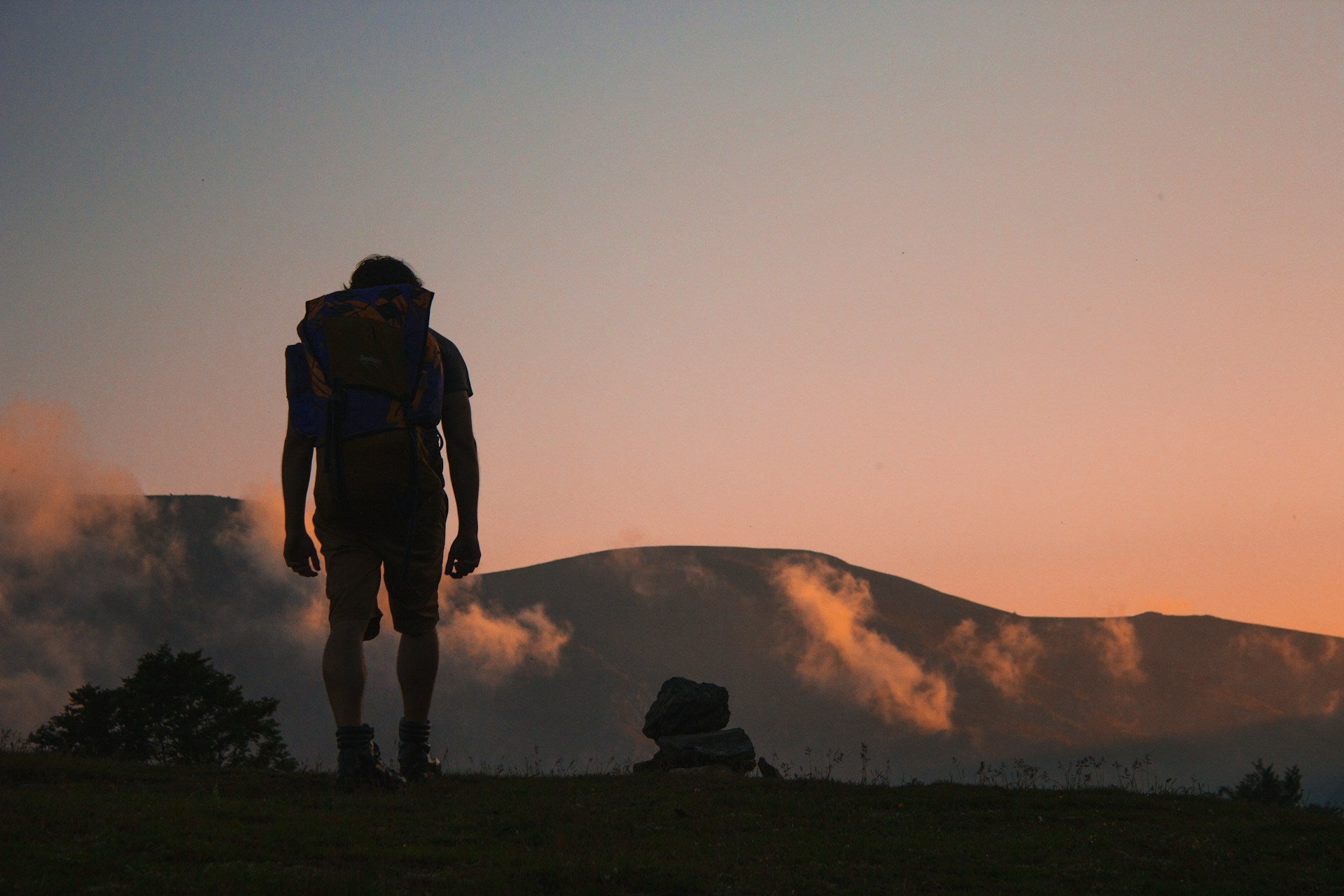 A hiker with a stuffed pack standing against a sunset over a mountain.