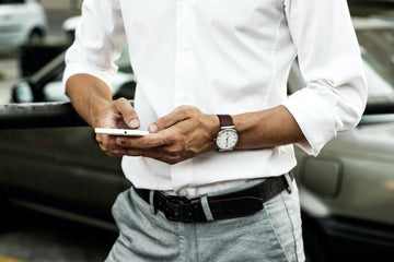 Man in white button up and light wash jeans with a black belt looks at his phone.